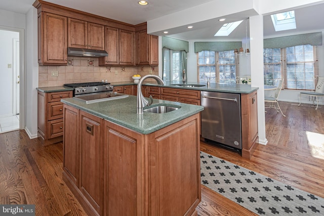 kitchen with brown cabinetry, a skylight, a sink, under cabinet range hood, and stainless steel dishwasher