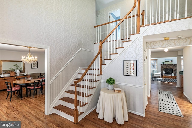 stairs with wallpapered walls, a wainscoted wall, a stone fireplace, wood finished floors, and a notable chandelier