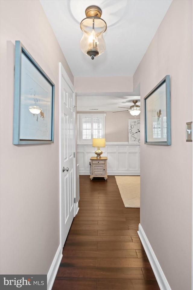 hallway featuring dark wood-style floors, a decorative wall, baseboards, and a wainscoted wall