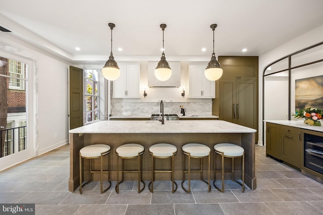 kitchen featuring a breakfast bar, an island with sink, decorative backsplash, wine cooler, and white cabinetry