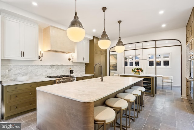 kitchen featuring an island with sink, a sink, custom range hood, white cabinetry, and tasteful backsplash