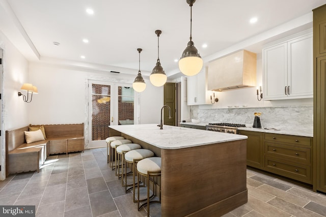 kitchen featuring custom exhaust hood, a kitchen island with sink, white cabinets, a kitchen bar, and backsplash