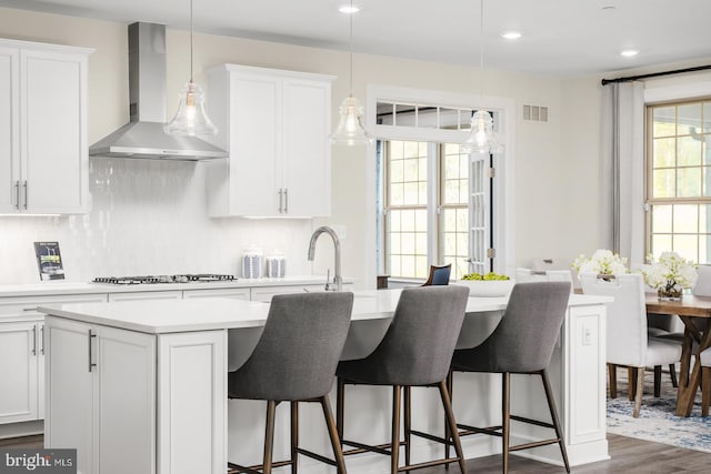 kitchen with dark wood-type flooring, a center island with sink, light countertops, white cabinetry, and wall chimney exhaust hood