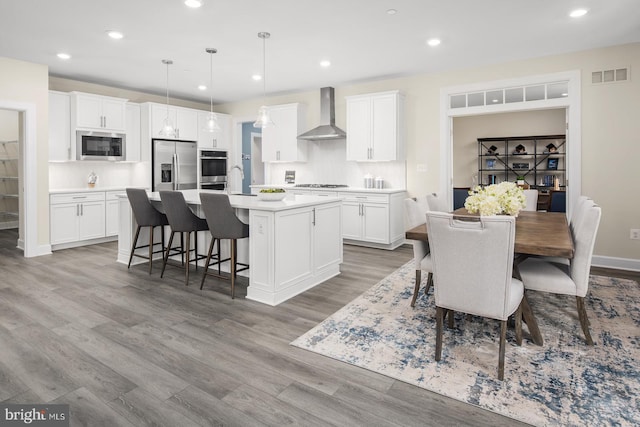 kitchen featuring visible vents, a kitchen island with sink, wood finished floors, stainless steel appliances, and wall chimney exhaust hood