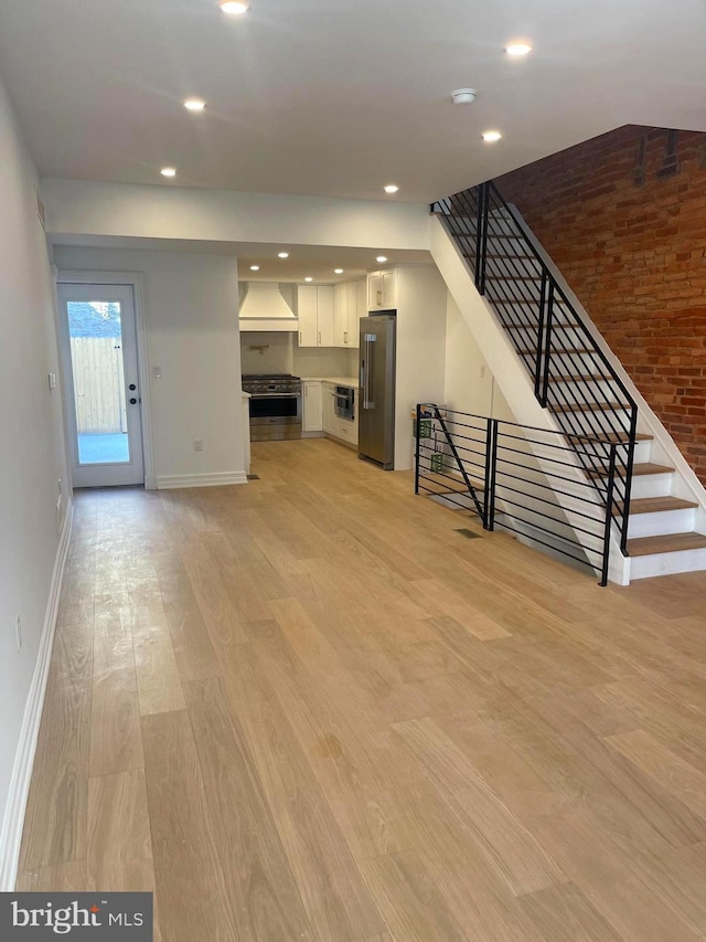 unfurnished living room featuring light wood finished floors, brick wall, baseboards, stairs, and recessed lighting