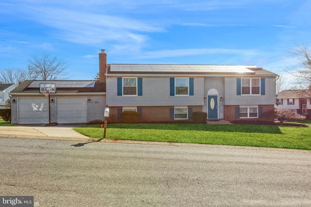 raised ranch featuring driveway, a front lawn, an attached garage, solar panels, and a chimney