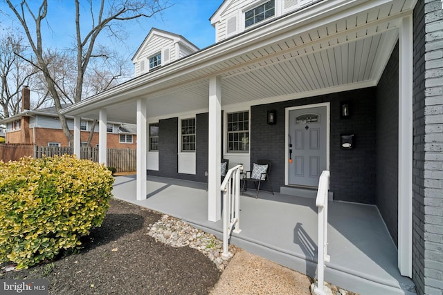 view of exterior entry with brick siding, covered porch, and fence