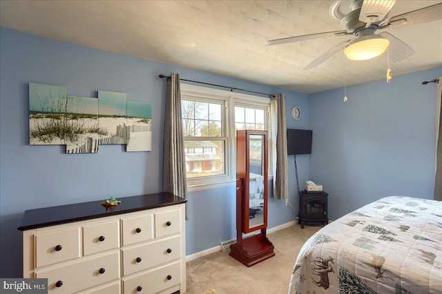 bedroom featuring light colored carpet, a ceiling fan, a wood stove, and baseboards