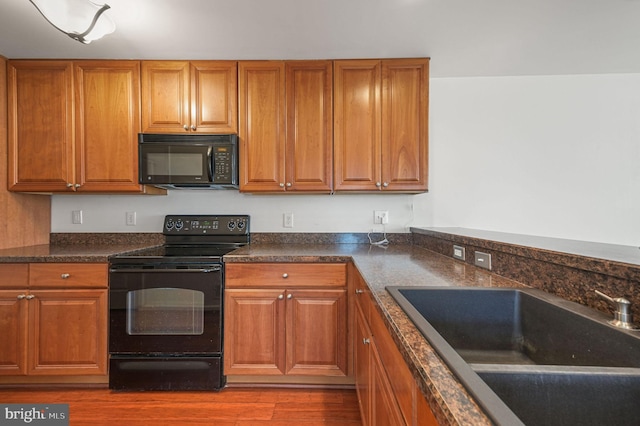 kitchen with black appliances, a sink, dark countertops, wood finished floors, and brown cabinetry