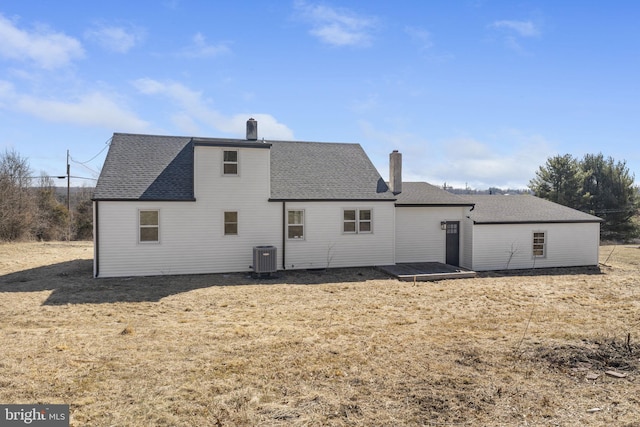 rear view of house featuring a yard, a chimney, central AC, and a shingled roof