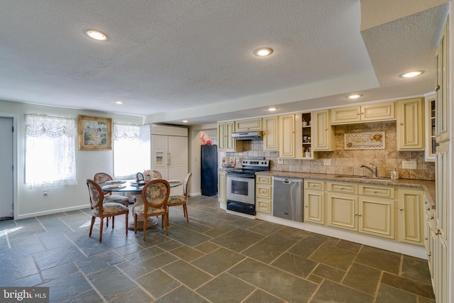 kitchen featuring a sink, under cabinet range hood, backsplash, cream cabinets, and appliances with stainless steel finishes