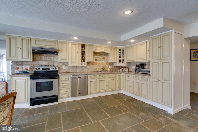 kitchen with under cabinet range hood, cream cabinets, appliances with stainless steel finishes, and stone tile floors