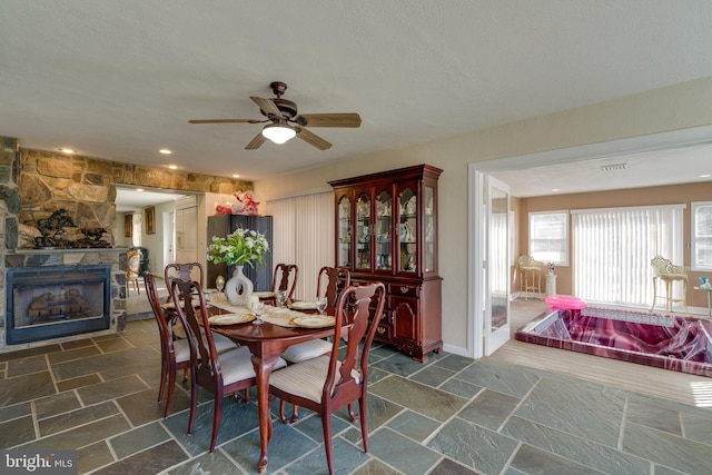 dining room featuring stone tile flooring, a fireplace, baseboards, and ceiling fan