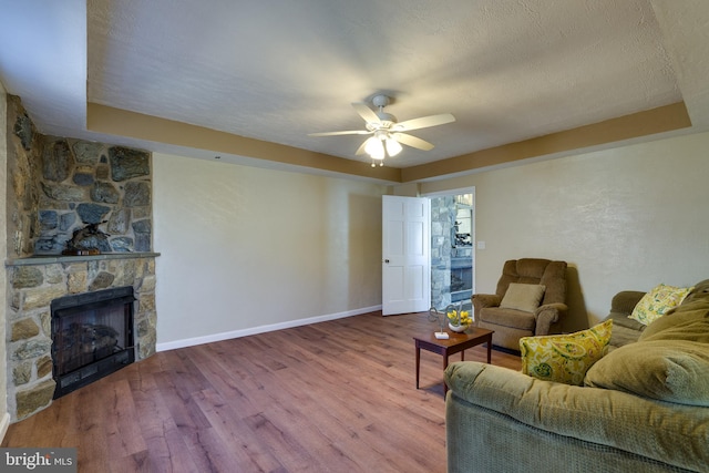 living area featuring a textured ceiling, wood finished floors, a stone fireplace, baseboards, and ceiling fan