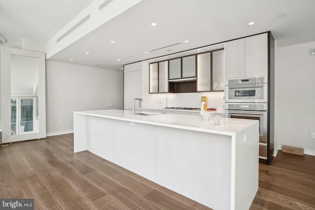 kitchen featuring a spacious island, dark wood-type flooring, light countertops, stainless steel double oven, and a sink