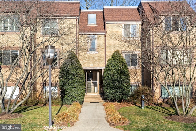 view of property featuring brick siding, mansard roof, a shingled roof, and a front lawn