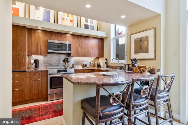 kitchen featuring a kitchen bar, appliances with stainless steel finishes, brown cabinetry, stone countertops, and a sink