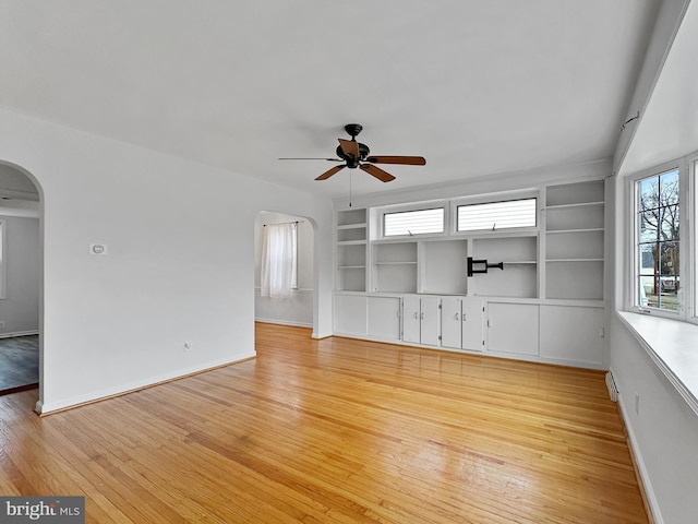 unfurnished living room with built in shelves, baseboards, ceiling fan, light wood-style floors, and arched walkways