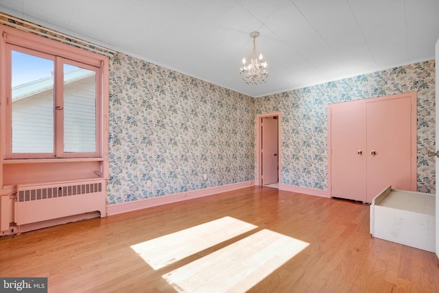 empty room with a notable chandelier, wallpapered walls, radiator, light wood finished floors, and baseboards