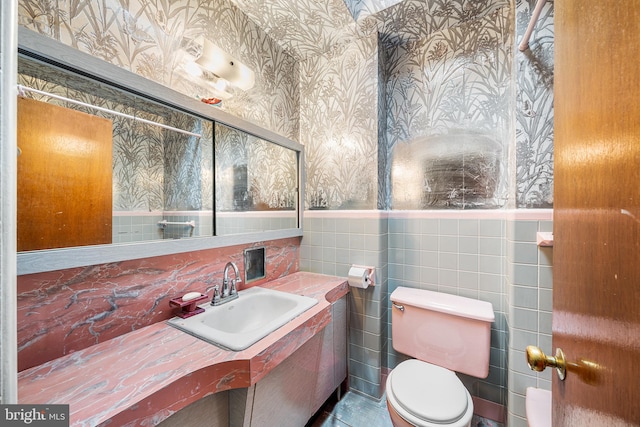 bathroom featuring wallpapered walls, a sink, wainscoting, tile walls, and toilet