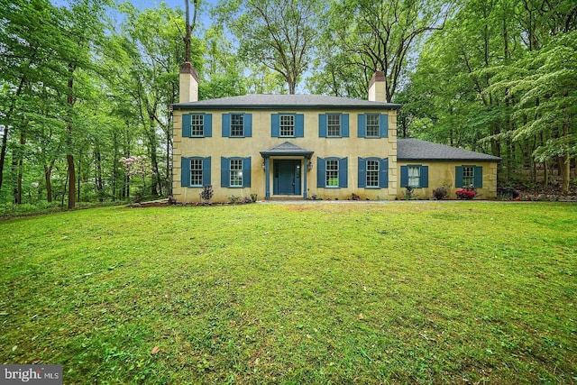 colonial home with stucco siding, a chimney, a front lawn, and a shingled roof