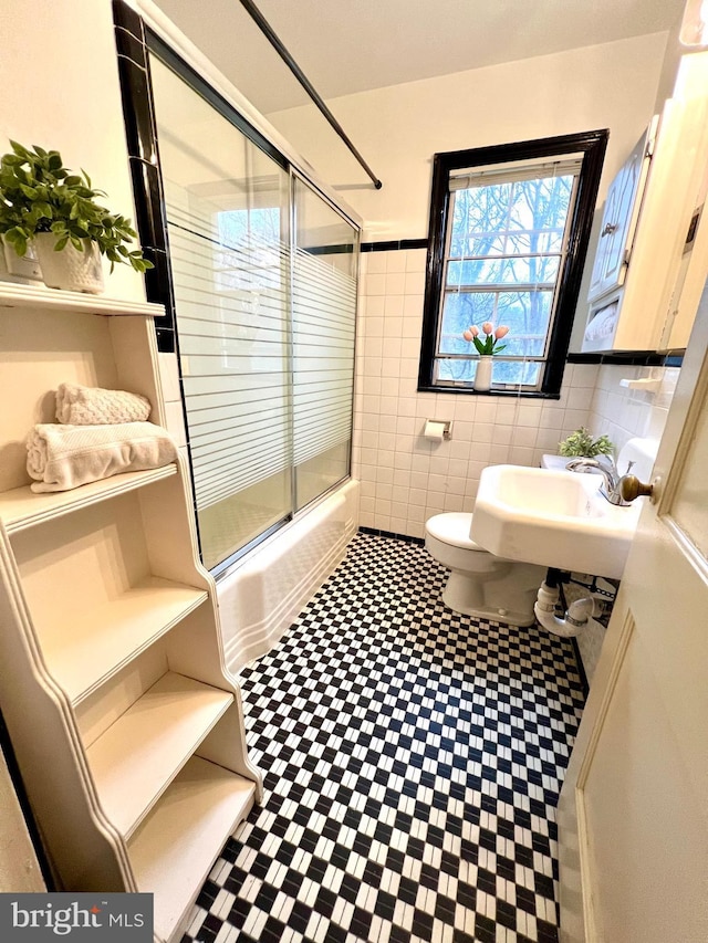 bathroom with a wainscoted wall, toilet, enclosed tub / shower combo, tile walls, and a sink