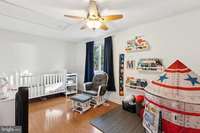 bedroom featuring a crib, wood finished floors, baseboards, attic access, and ceiling fan