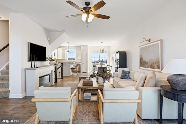 living area featuring baseboards, stairway, ceiling fan with notable chandelier, wood finished floors, and a glass covered fireplace