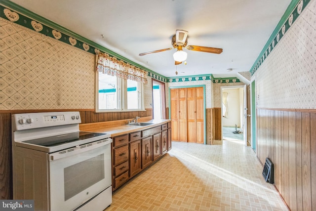 kitchen with a wainscoted wall, brick patterned floor, a ceiling fan, white electric range oven, and wallpapered walls