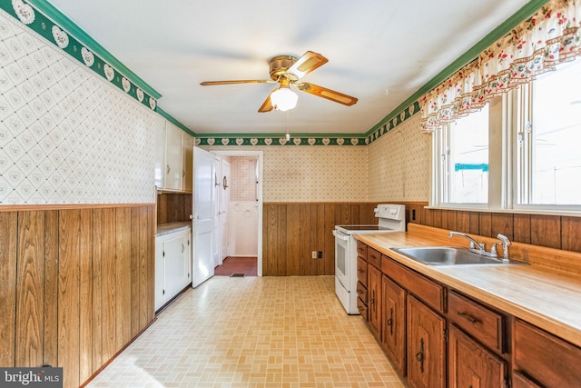 kitchen with a wainscoted wall, a ceiling fan, electric stove, a sink, and wallpapered walls