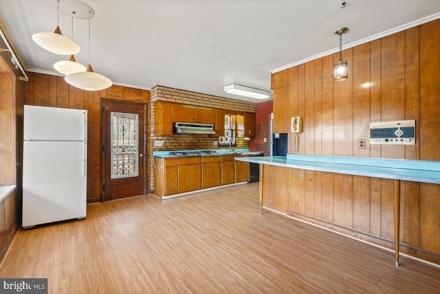 kitchen with under cabinet range hood, freestanding refrigerator, light wood-style floors, stainless steel gas stovetop, and brown cabinetry