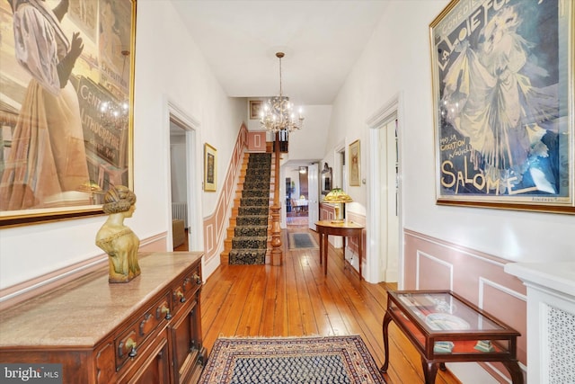 hall with an inviting chandelier, light wood-style flooring, stairway, and a wainscoted wall