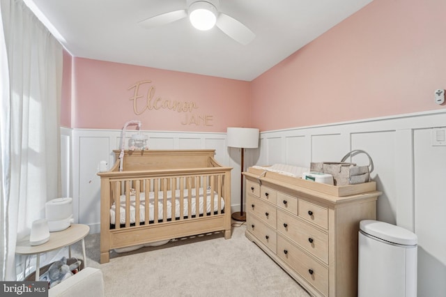 carpeted bedroom with wainscoting, a crib, and a ceiling fan