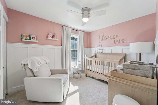 bedroom with a crib, a ceiling fan, carpet floors, and wainscoting