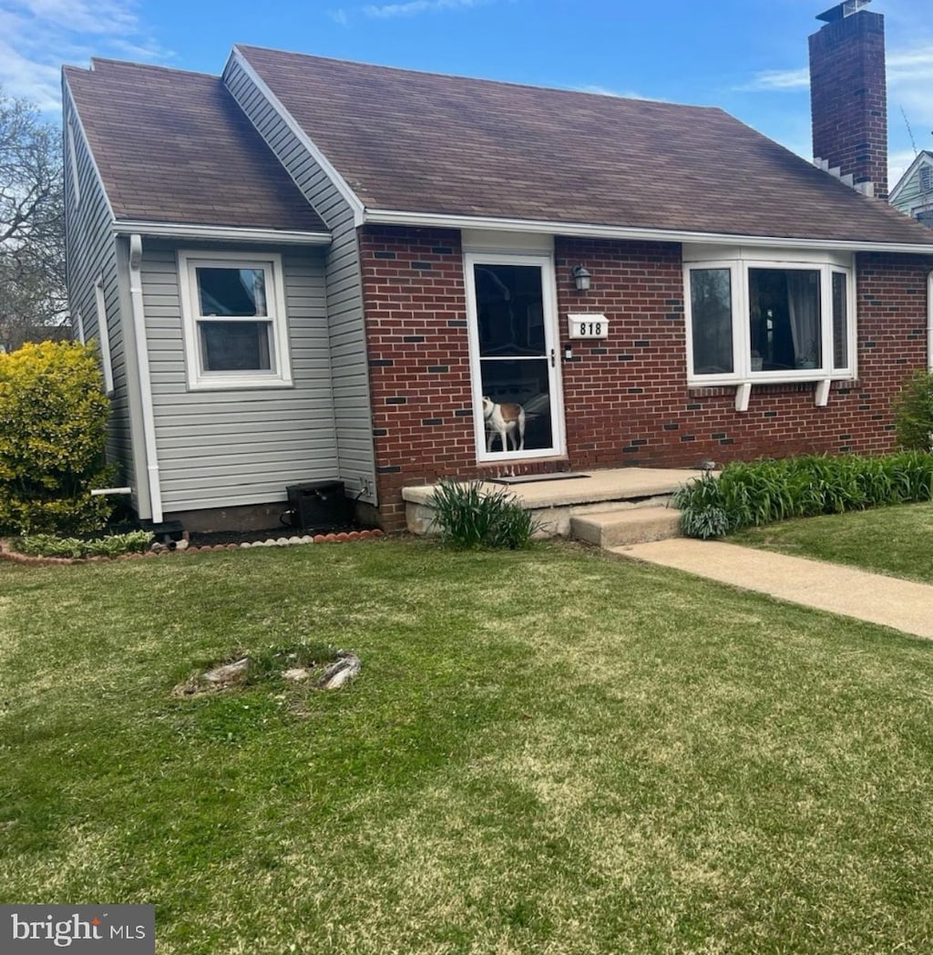 view of front facade with a shingled roof, a front yard, brick siding, and a chimney