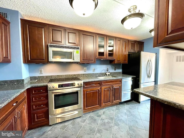 kitchen featuring glass insert cabinets, dark stone countertops, stainless steel appliances, a textured ceiling, and a sink