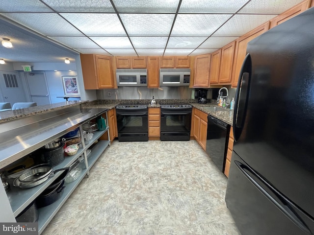 kitchen featuring dark stone counters, brown cabinets, black appliances, a paneled ceiling, and a sink