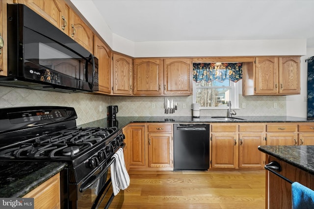 kitchen featuring light wood finished floors, dark stone countertops, brown cabinetry, black appliances, and a sink