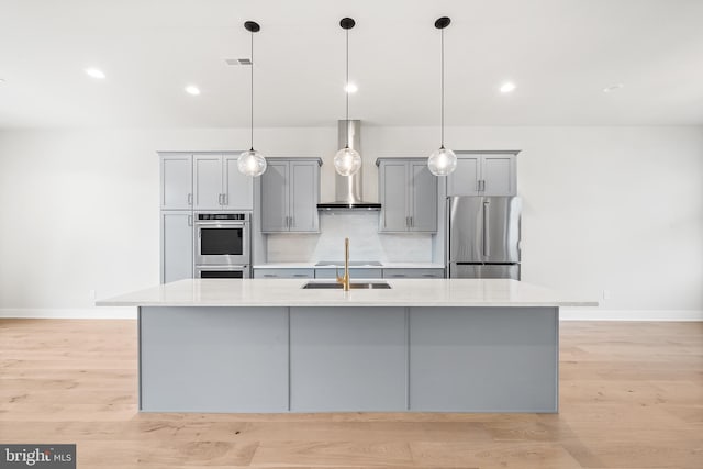 kitchen featuring an island with sink, gray cabinets, stainless steel appliances, wall chimney exhaust hood, and light countertops