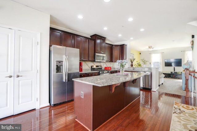 kitchen with dark brown cabinets, backsplash, a center island, light stone countertops, and stainless steel appliances