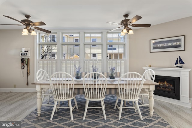 dining space featuring wood finished floors, baseboards, a warm lit fireplace, and ceiling fan