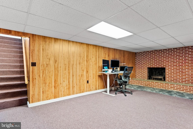 unfurnished office with a brick fireplace, baseboards, wood walls, carpet floors, and a paneled ceiling