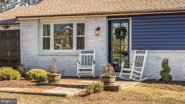 view of exterior entry with an attached garage, brick siding, and roof with shingles