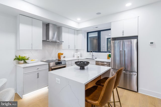 kitchen featuring wall chimney range hood, a kitchen island, a sink, appliances with stainless steel finishes, and a kitchen breakfast bar