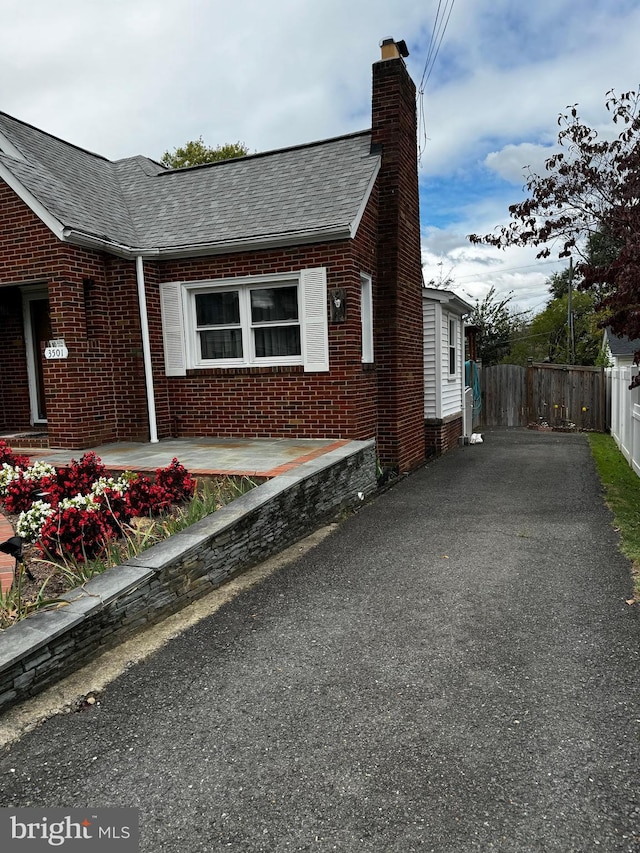 view of side of home with fence, roof with shingles, a chimney, aphalt driveway, and brick siding