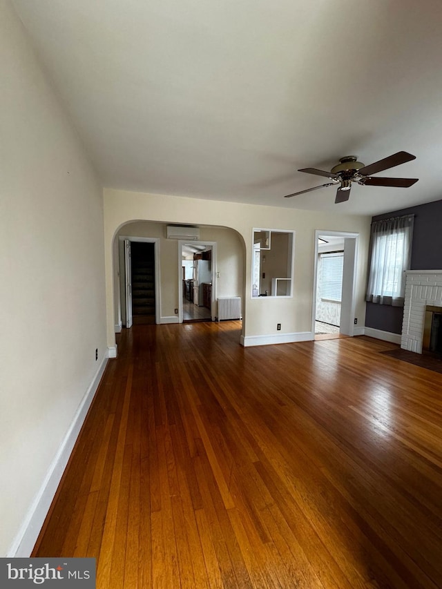 unfurnished living room featuring arched walkways, radiator, wood-type flooring, baseboards, and ceiling fan