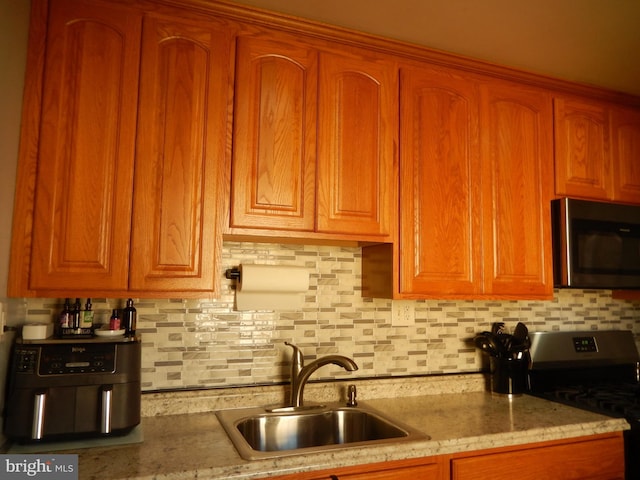 kitchen with light stone counters, decorative backsplash, brown cabinets, stainless steel appliances, and a sink