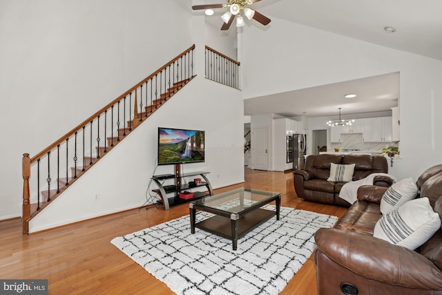 living area with baseboards, stairway, ceiling fan with notable chandelier, light wood-style floors, and high vaulted ceiling