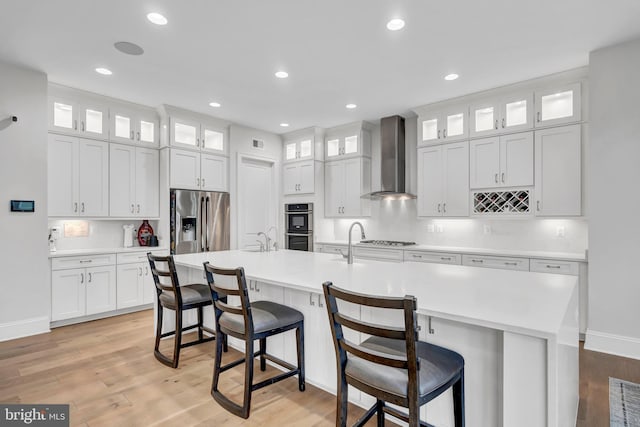 kitchen with wall chimney range hood, an island with sink, light wood-type flooring, and stainless steel appliances