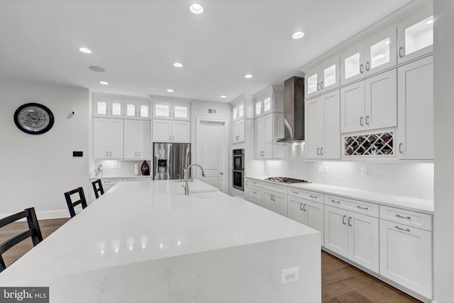 kitchen featuring a large island with sink, appliances with stainless steel finishes, white cabinets, wall chimney exhaust hood, and a sink